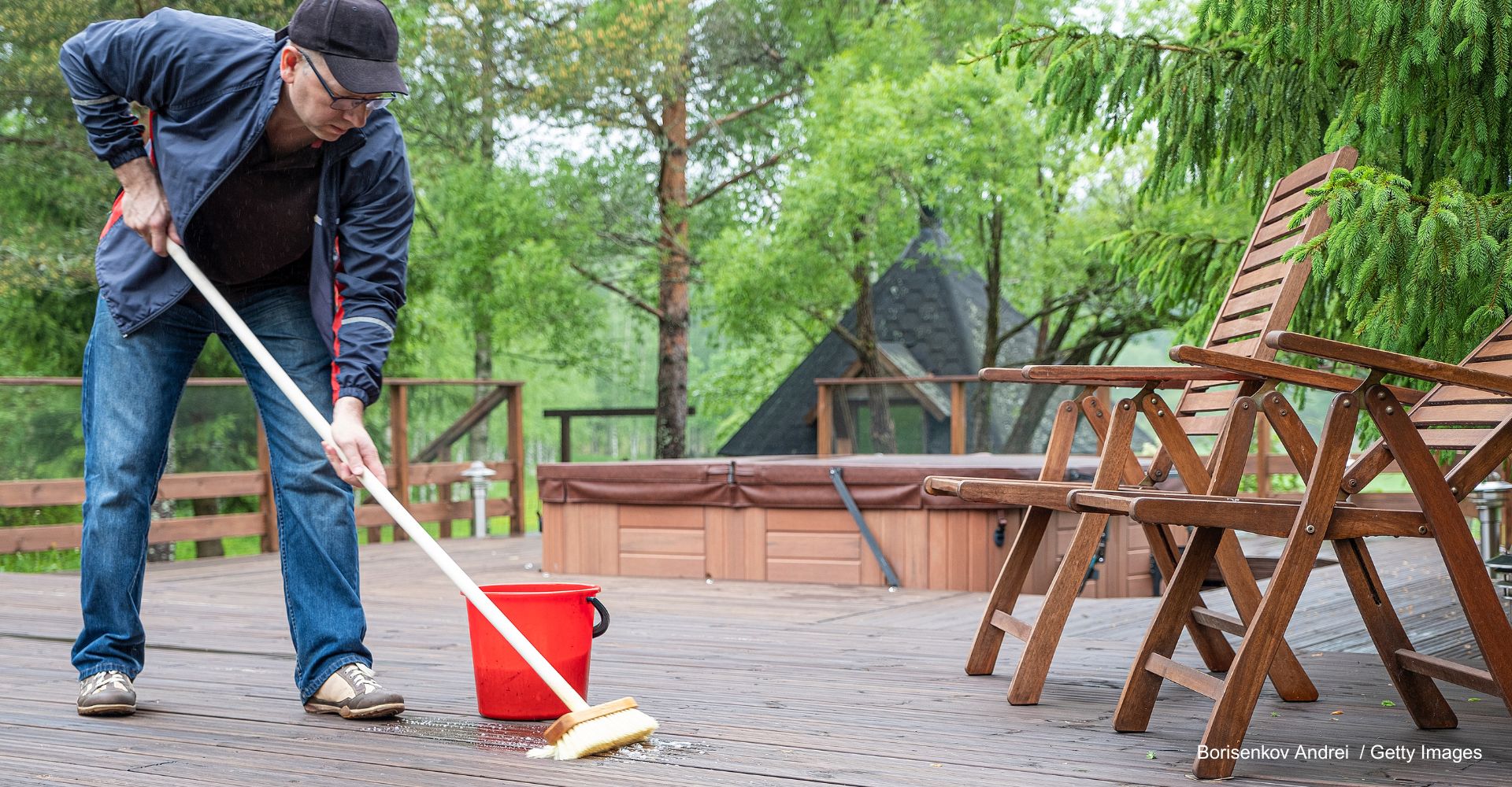 Mann mit Jacke und Hut reinigt Terrassendielen mit Wasser, milder Seife und Schrubber, neben ihm steht ein roter Eimer